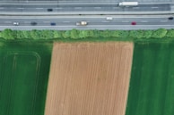 Aerial view of a highway with vehicles traveling in both directions, adjacent to a field. The field has distinct sections where one is freshly plowed and others are lush green. A line of trees provides a natural barrier between the highway and the agricultural land.