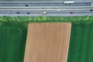 Aerial view of a highway with vehicles traveling in both directions, adjacent to a field. The field has distinct sections where one is freshly plowed and others are lush green. A line of trees provides a natural barrier between the highway and the agricultural land.