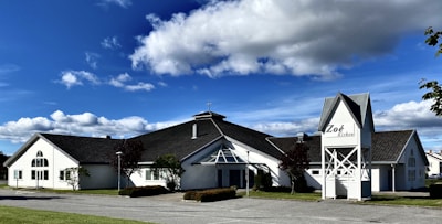A white building with a cross on the roof, featuring modern architectural elements and large windows. It is surrounded by well-maintained greenery, with a bright blue sky and scattered clouds in the background. The sign on the building reads 'Zoé Kirken.'