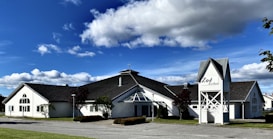 A white building with a cross on the roof, featuring modern architectural elements and large windows. It is surrounded by well-maintained greenery, with a bright blue sky and scattered clouds in the background. The sign on the building reads 'Zo&eacute; Kirken.'
