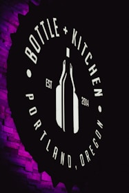 A circular sign features the words 'Bottle + Kitchen' and 'Portland, Oregon' with an outline of a wine bottle in the center. The background is dark, and the sign is illuminated, creating a contrast with the surrounding purple-lit textured wall.