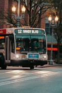 A public transport bus displaying the route number 12 and the destination 'Sandy Blvd' is captured in motion. The bus is part of the TriMet transportation service, evident from the logo on the front. Streetlights illuminate the scene, and there are trees and buildings in the background. The scene depicts an urban setting.