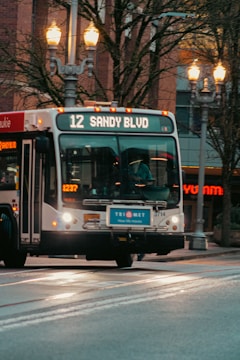 A public transport bus displaying the route number 12 and the destination 'Sandy Blvd' is captured in motion. The bus is part of the TriMet transportation service, evident from the logo on the front. Streetlights illuminate the scene, and there are trees and buildings in the background. The scene depicts an urban setting.