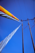 A close-up view of electrical transmission lines stretching into the distance against a clear blue sky. The perspective emphasizes the spiraled metal wires with a yellow and orange protective covering. Tall poles support the wires, and the composition highlights the lines converging in the distance.