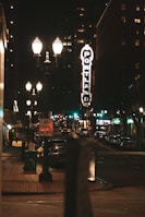 A nighttime urban street scene with vintage-style street lamps illuminating the sidewalk and street. A prominent vertical sign displays the word 'Portland' in bright lights. Buildings line the street, and several vehicles are parked along the roadside. A 'No Turn On Red' traffic sign is visible in the foreground.