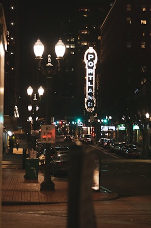 A nighttime urban street scene with vintage-style street lamps illuminating the sidewalk and street. A prominent vertical sign displays the word 'Portland' in bright lights. Buildings line the street, and several vehicles are parked along the roadside. A 'No Turn On Red' traffic sign is visible in the foreground.