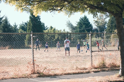 Children are playing soccer on a grassy field surrounded by a chain-link fence. A coach or adult stands nearby watching the game. Trees and greenery are visible in the background, and the scene is lit by daytime sunlight.