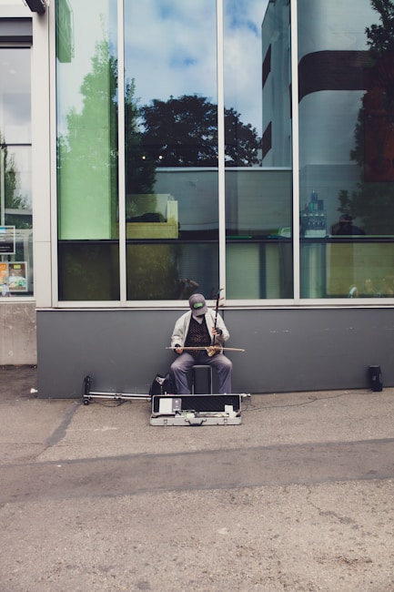 A street musician is seated against a large glass window, playing a string instrument. The musician wears casual clothing and a cap, and his equipment is placed on the ground in front of him. The background features reflections of trees and the sky in the glass.