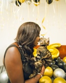 A person is holding a cat while their noses touch affectionately amidst a background of gold, silver, and black balloons. The setting suggests a celebratory atmosphere, possibly a party or special event.