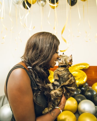 A person is holding a cat while their noses touch affectionately amidst a background of gold, silver, and black balloons. The setting suggests a celebratory atmosphere, possibly a party or special event.