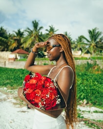 A woman holding a bouquet of vibrant red roses with lush greenery in the background and palm trees under a bright sky. She is wearing sunglasses and has long braided hair, shielding her eyes from the sunlight.