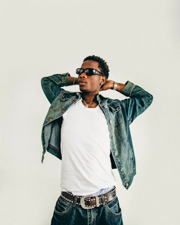 A person wearing sunglasses and denim clothing poses confidently with their hands behind their head. The individual wears a white shirt underneath a denim jacket, a studded belt, and denim jeans.