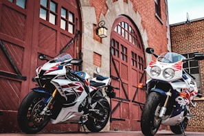 Two sporty motorcycles are parked in front of a historical brick building with large wooden doors. The motorcycles have sleek designs with red, white, and blue color schemes, featuring brand logos. The scene captures a mix of modern technology and classic architecture.