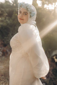 A person is wearing a flowing white gown adorned with a floral headpiece featuring pale blue flowers. The background is softly lit with sunlight filtering through surrounding greenery, creating a serene and gentle atmosphere.