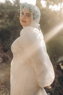 A person is wearing a flowing white gown adorned with a floral headpiece featuring pale blue flowers. The background is softly lit with sunlight filtering through surrounding greenery, creating a serene and gentle atmosphere.