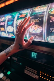 A hand with tattoos reaches towards a brightly lit slot machine, featuring various colorful symbols including a tiger. The image captures the moment of anticipation as the person engages with the machine during a game of chance.