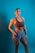 A person stands confidently against a solid turquoise background, wearing a brown sports bra and gray-blue athletic shorts. The individual has a fit physique and is adorned with earrings and a bracelet. Tattoos are visible on their arm, adding to their sporty appearance.