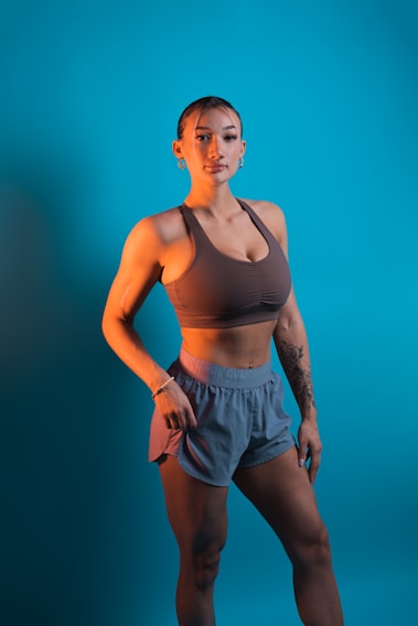 A person stands confidently against a solid turquoise background, wearing a brown sports bra and gray-blue athletic shorts. The individual has a fit physique and is adorned with earrings and a bracelet. Tattoos are visible on their arm, adding to their sporty appearance.
