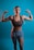 A woman with braided hair poses confidently with both arms flexed against a blue background. She is wearing a brown sports bra and gray shorts, showcasing her toned muscles. The lighting emphasizes her physique and creates a strong, empowering image.