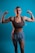 A woman with braided hair poses confidently with both arms flexed against a blue background. She is wearing a brown sports bra and gray shorts, showcasing her toned muscles. The lighting emphasizes her physique and creates a strong, empowering image.