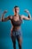 A woman with braided hair poses confidently with both arms flexed against a blue background. She is wearing a brown sports bra and gray shorts, showcasing her toned muscles. The lighting emphasizes her physique and creates a strong, empowering image.