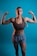 A woman with braided hair poses confidently with both arms flexed against a blue background. She is wearing a brown sports bra and gray shorts, showcasing her toned muscles. The lighting emphasizes her physique and creates a strong, empowering image.