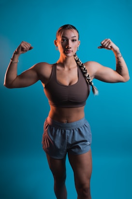 A woman with braided hair poses confidently with both arms flexed against a blue background. She is wearing a brown sports bra and gray shorts, showcasing her toned muscles. The lighting emphasizes her physique and creates a strong, empowering image.