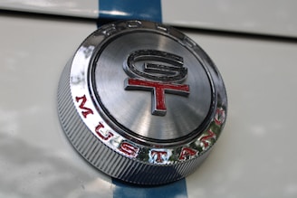 A close-up view of a metallic emblem with 'GT' inscribed in the center and 'MUSTANG' written around the bottom. The emblem is circular with a shiny silver finish, featuring red and black text, and is set against a white surface with a blue stripe.