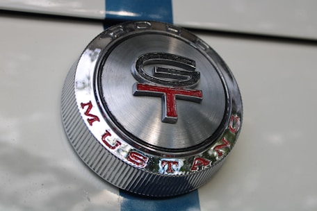 A close-up view of a metallic emblem with 'GT' inscribed in the center and 'MUSTANG' written around the bottom. The emblem is circular with a shiny silver finish, featuring red and black text, and is set against a white surface with a blue stripe.