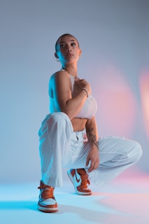 A person poses confidently in a studio setting with a gradient blue and pink backdrop. They are wearing a white sleeveless top, white pants, and orange and white sneakers. The lighting creates a dynamic effect with contrasting colors highlighting different aspects of the scene.