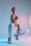 A person poses confidently in a studio setting with a gradient blue and pink backdrop. They are wearing a white sleeveless top, white pants, and orange and white sneakers. The lighting creates a dynamic effect with contrasting colors highlighting different aspects of the scene.