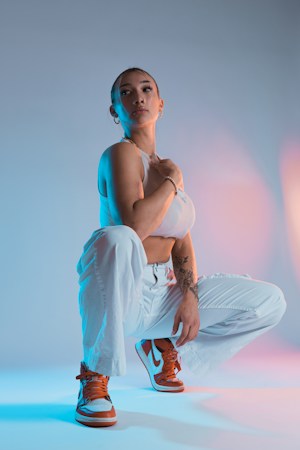 A person poses confidently in a studio setting with a gradient blue and pink backdrop. They are wearing a white sleeveless top, white pants, and orange and white sneakers. The lighting creates a dynamic effect with contrasting colors highlighting different aspects of the scene.