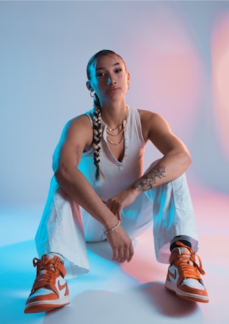 A person with a braided hairstyle is sitting in a relaxed pose on the floor, wearing a white sleeveless top, white pants, and orange-and-white sneakers. Multiple necklaces adorn her neck and her tattoos are visible on her right arm. The background is lit with a gradient of blue and pink lighting, creating a vibrant and dynamic contrast.