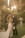 A couple is posing for a wedding photograph in an outdoor garden setting. The bride is in a white gown with floral details, holding a bouquet of white flowers. The groom is wearing a dark suit with a white boutonniere. They are surrounded by lush greenery and soft, warm sunlight filters through from above, creating a dreamy atmosphere.