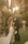 A couple is posing for a wedding photograph in an outdoor garden setting. The bride is in a white gown with floral details, holding a bouquet of white flowers. The groom is wearing a dark suit with a white boutonniere. They are surrounded by lush greenery and soft, warm sunlight filters through from above, creating a dreamy atmosphere.