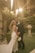 A couple is posing for a wedding photograph in an outdoor garden setting. The bride is in a white gown with floral details, holding a bouquet of white flowers. The groom is wearing a dark suit with a white boutonniere. They are surrounded by lush greenery and soft, warm sunlight filters through from above, creating a dreamy atmosphere.