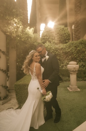 A couple is posing for a wedding photograph in an outdoor garden setting. The bride is in a white gown with floral details, holding a bouquet of white flowers. The groom is wearing a dark suit with a white boutonniere. They are surrounded by lush greenery and soft, warm sunlight filters through from above, creating a dreamy atmosphere.
