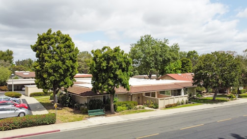 A residential area with single-story houses surrounded by large trees. The houses have brown roofs and light-colored walls. A street runs in front of the houses, with a sidewalk and a few parked cars visible. A green bench is placed on the grass between the sidewalk and the houses. The sky is partly cloudy.