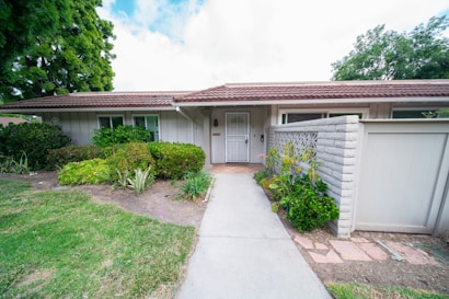 A single-story house with a white exterior, brown roof tiles, and surrounded by lush green plants. There is a concrete path leading to the front door, flanked by well-maintained shrubs and a small garden area. A white fence and a brick wall are part of the property&rsquo;s boundary, with tall trees visible in the background.