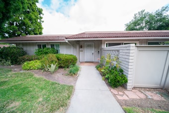 A single-story house with a white exterior, brown roof tiles, and surrounded by lush green plants. There is a concrete path leading to the front door, flanked by well-maintained shrubs and a small garden area. A white fence and a brick wall are part of the property’s boundary, with tall trees visible in the background.