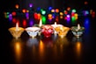 A set of ornately decorated clay lamps are lit, casting a warm glow on a dark surface. In the background, colorful and blurred string lights of various colors create a festive ambiance.