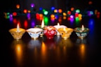A set of ornately decorated clay lamps are lit, casting a warm glow on a dark surface. In the background, colorful and blurred string lights of various colors create a festive ambiance.