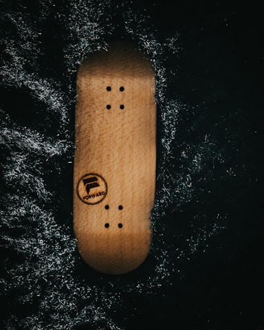 A wooden skateboard deck with screw holes is placed against a dark, textured background. The word 'FORWARD' and a logo are visible on the deck.