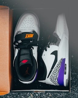 A pair of stylish white and black athletic shoes with orange and red branding, featuring purple and elephant print accents, placed inside a cardboard shoe box on a dark surface.