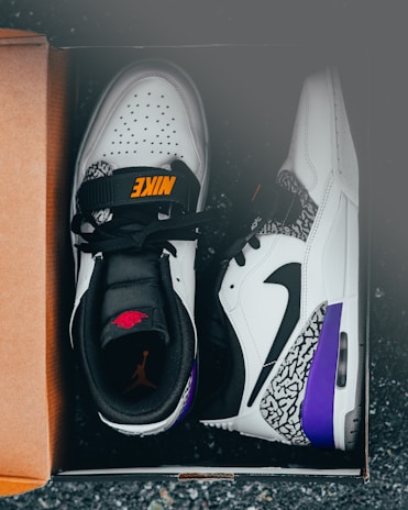A pair of stylish white and black athletic shoes with orange and red branding, featuring purple and elephant print accents, placed inside a cardboard shoe box on a dark surface.