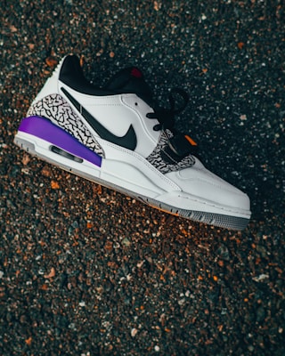 A stylish sneaker with a black swoosh and a mix of white leather and patterned fabric. The shoe features purple accents and is placed on a textured asphalt surface.