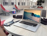A laptop is placed on a round table in a modern, bright indoor environment with large windows. The table also has a smartphone, a pair of headphones, a bottle of Pepsi, and some other items. The background includes stylish, modern furniture and green plants mounted on the wall.
