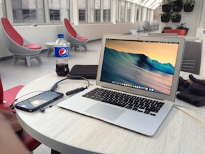 A laptop is placed on a round table in a modern, bright indoor environment with large windows. The table also has a smartphone, a pair of headphones, a bottle of Pepsi, and some other items. The background includes stylish, modern furniture and green plants mounted on the wall.
