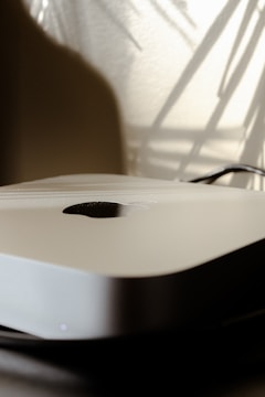 A close-up of a sleek electronic device with a metallic finish, featuring a logo in the center. The background shows soft, abstract shadows and lighting, suggesting a modern, minimalist aesthetic.