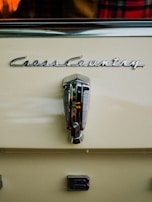 A close-up view of a vintage car with the logo 'Cross Country' prominently displayed above a metallic handle. The car is painted in a creamy beige color. There is a partial view of a plaid fabric and an orange object reflected in the background.